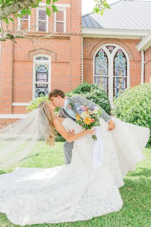 A flowing white wedding dress spreads across green grass in front of a red brick building.