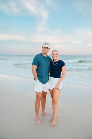 A couple stands barefoot on a sandy beach at sunset with ocean waves and blue sky in the background.