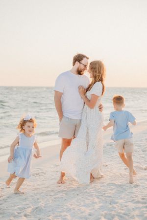 A family walks together along a white sandy beach at sunset wearing light blue and white outfits.