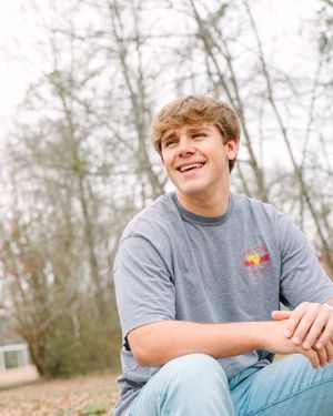 Young person in grey t-shirt sitting outdoors and smiling while relaxing in a natural wooded setting.