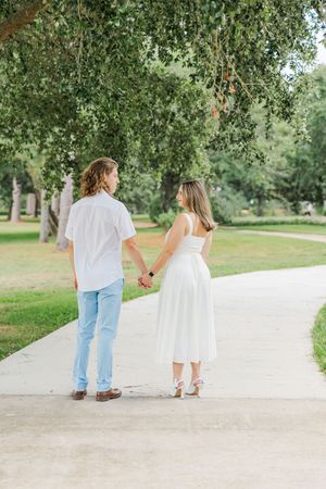 A couple holds hands walking along a park path, man in white shirt and jeans, woman in white midi dress.