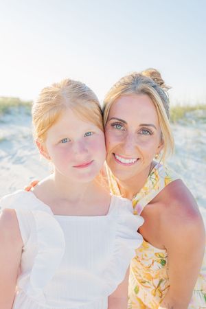A smiling blonde woman and young girl with blue eyes pose together on a sunny beach, wearing white and yellow floral outfits.