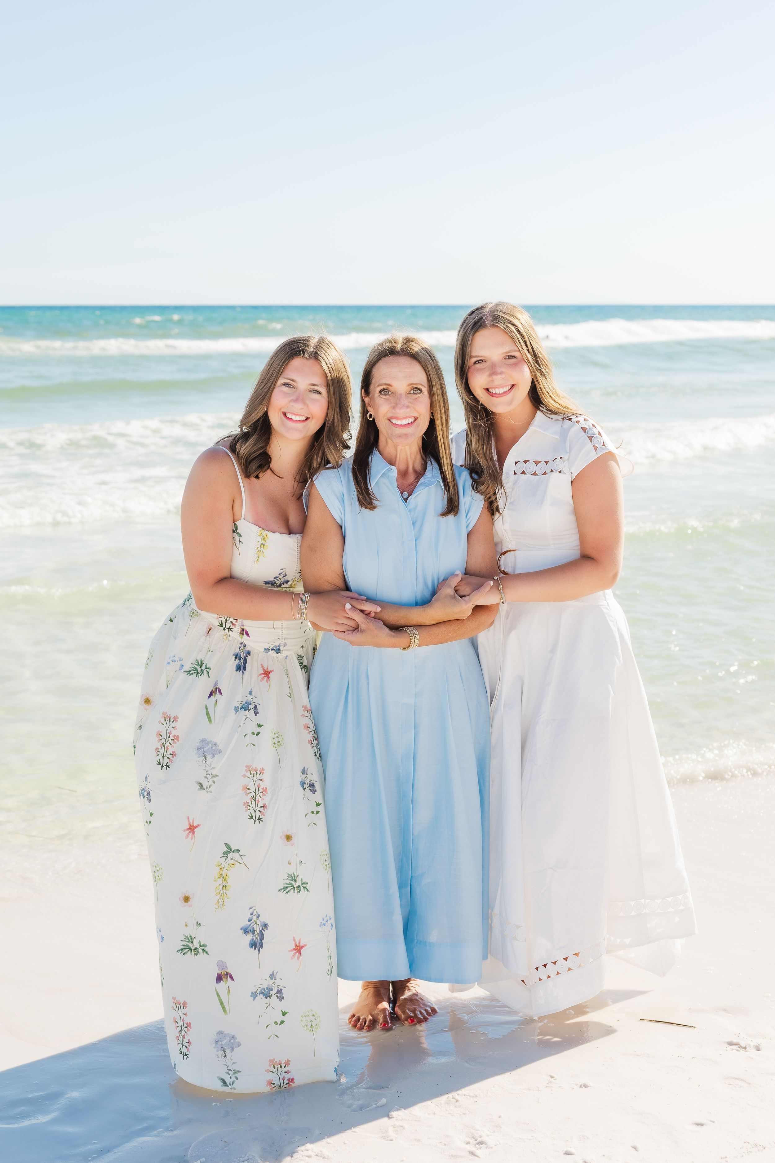 Three smiling women in light dresses pose together on a sunny sandy beach with turquoise ocean waves behind them.