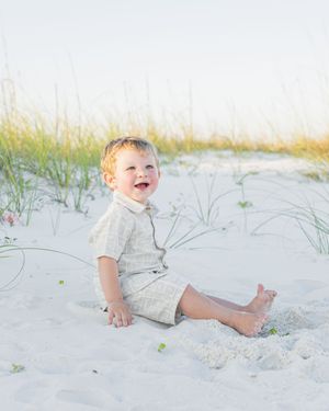 A happy toddler boy sits barefoot on white sand beach dunes, wearing a linen outfit, smiling joyfully outdoors.