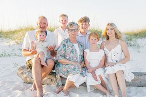 Multi-generational family of seven poses together on sandy beach, smiling warmly in light-colored outfits at sunset.
