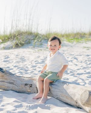 A smiling toddler in a white shirt and green shorts sits barefoot on driftwood at a sandy beach.