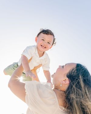 A joyful baby laughs while being lifted by a woman outdoors in bright sunlight.