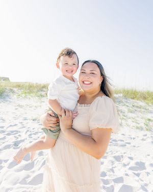 A smiling woman in a cream dress holds a happy toddler boy on a sunny white sand beach.