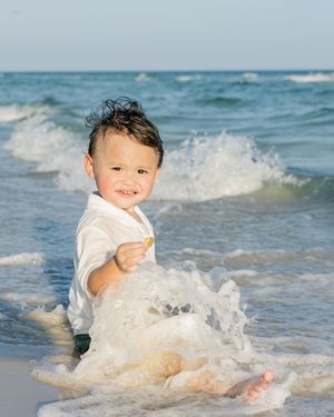 A happy toddler boy splashing in ocean waves on a sunny beach, wearing a white shirt.