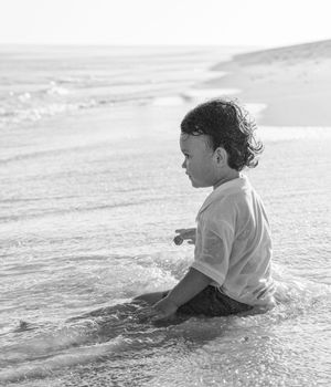 Black and white photo of a young child sitting in ocean waves on a sandy beach, wearing a white t-shirt.