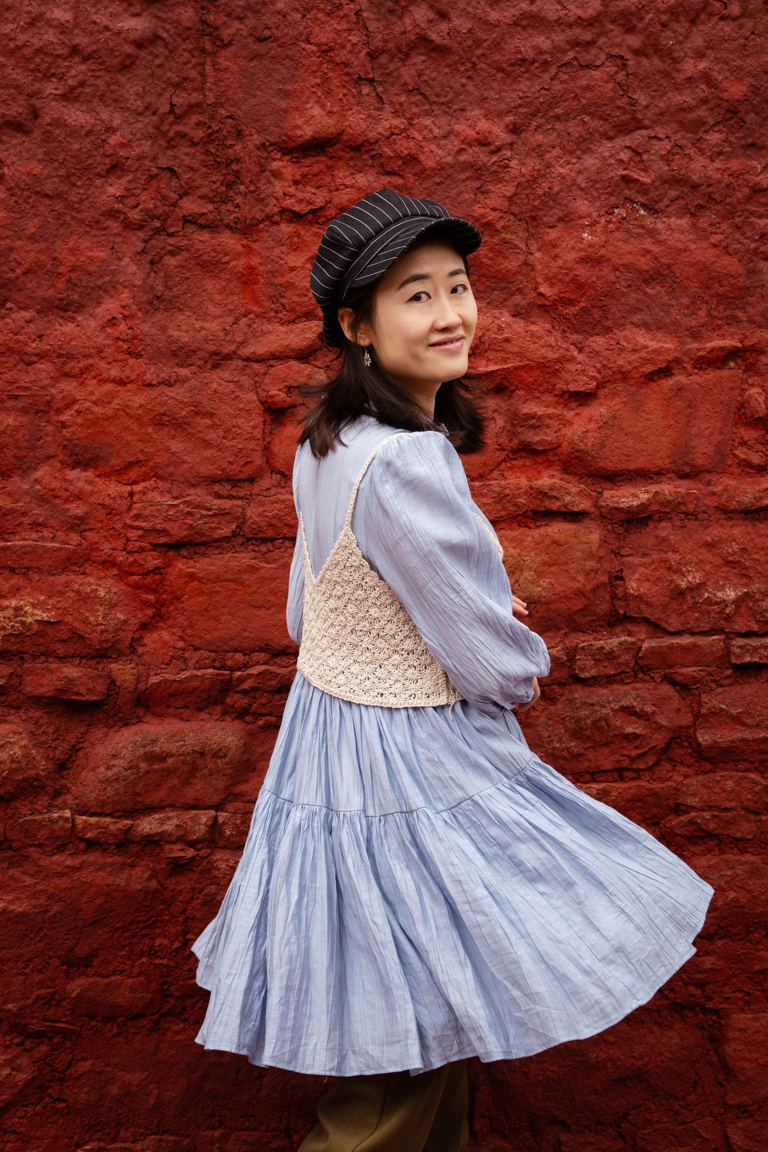 A playful pose against a red brick wall showcases a blue vintage-style dress and newsboy cap.