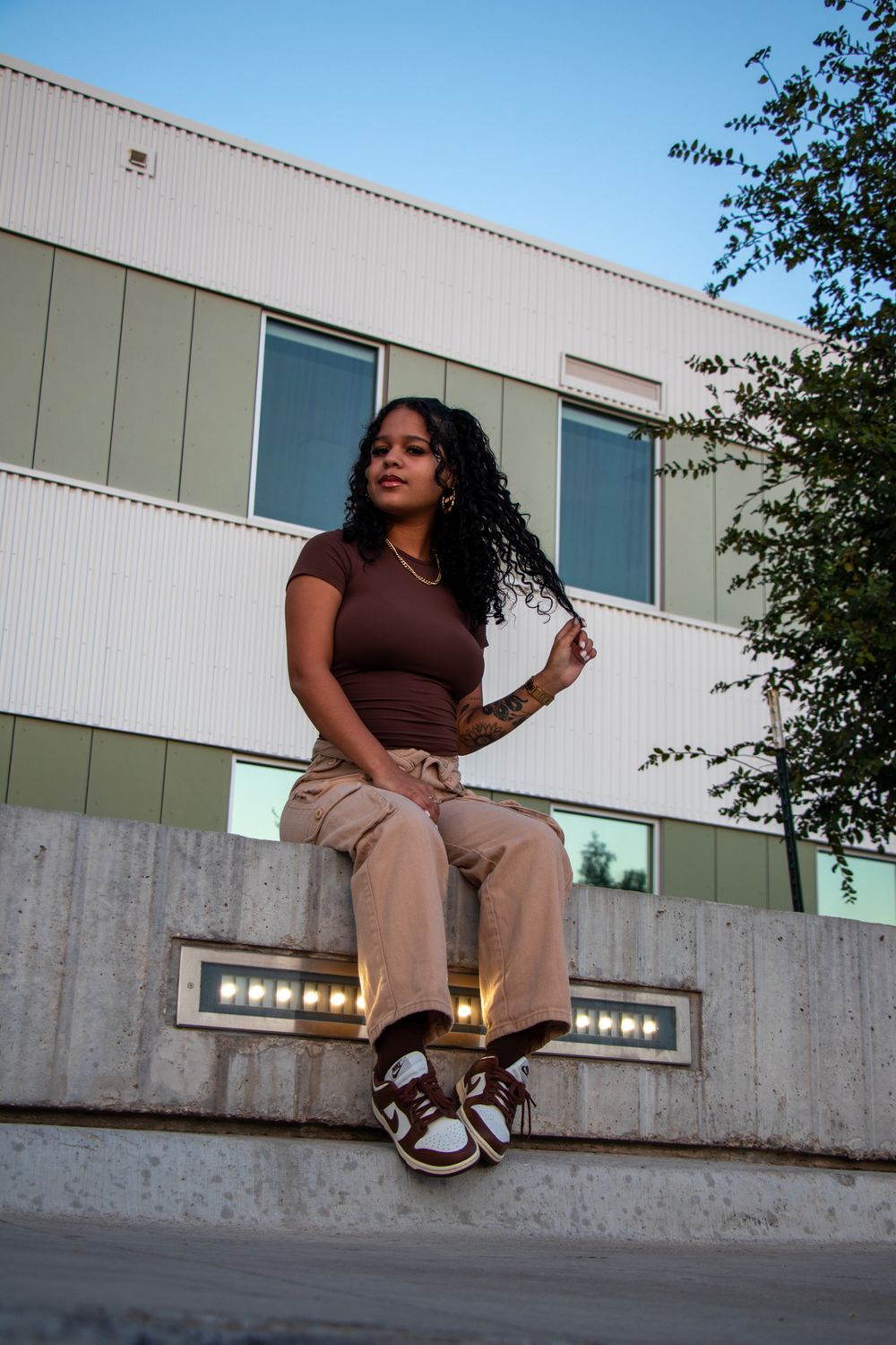 A person in brown top and beige pants sits on a concrete ledge against a modern building exterior.
