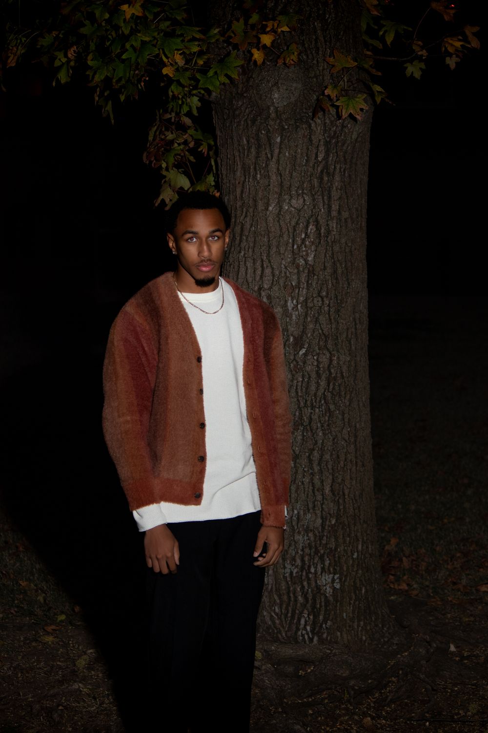 Person in brown cardigan and white shirt standing against dark tree backdrop at night.