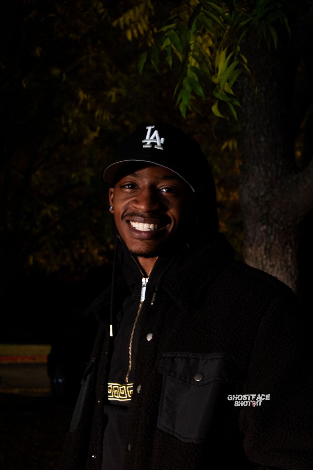 A person wearing a Los Angeles Dodgers cap and black jacket poses at night against a dark background with autumn foliage.