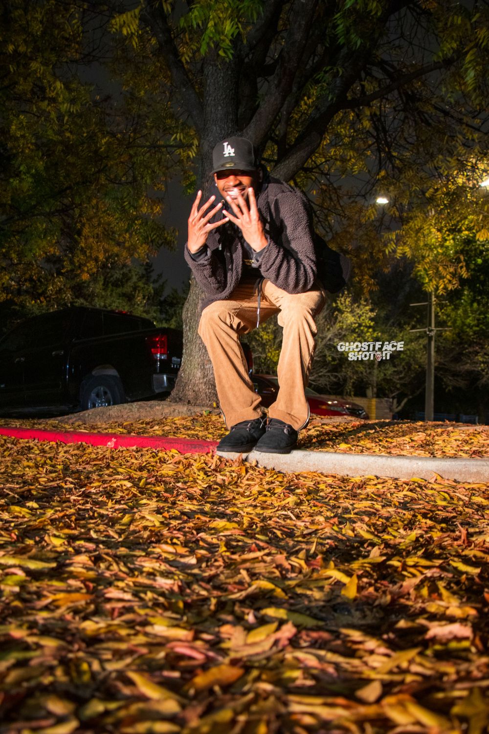 Someone in a baseball cap covers their face while sitting on a curb surrounded by autumn leaves.