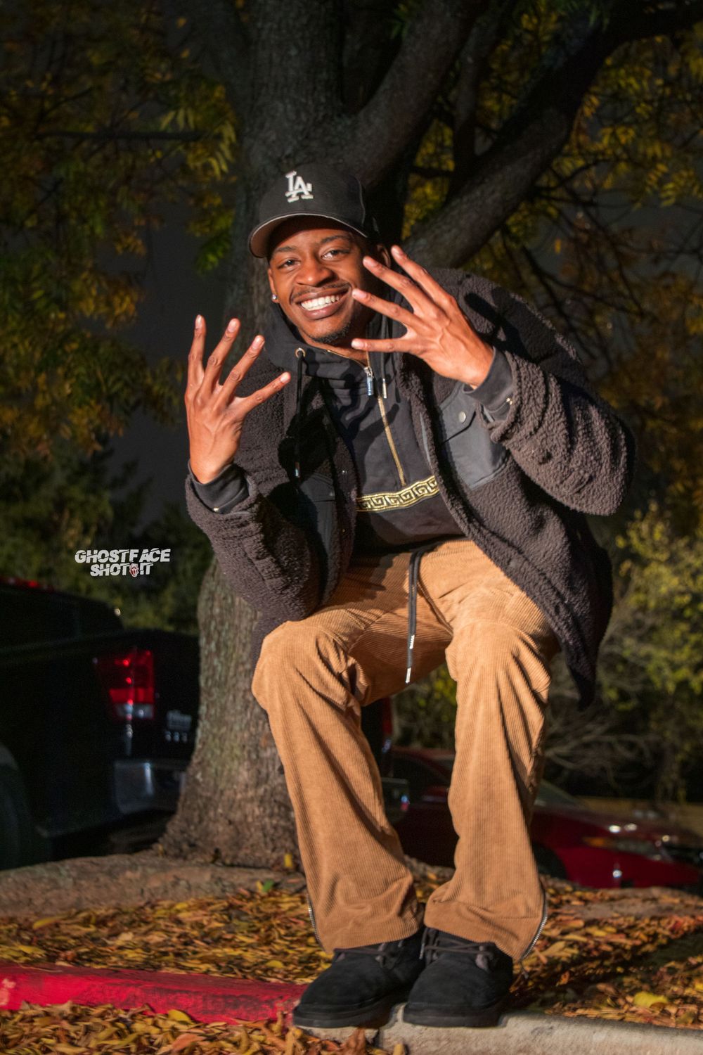 A figure in casual attire gestures playfully while sitting outdoors among fallen autumn leaves.
