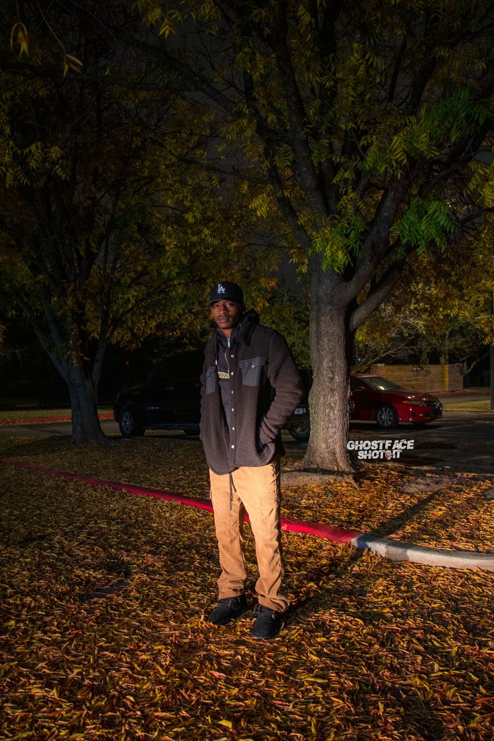 A casual portrait of someone standing on fallen leaves wearing khaki pants and a dark jacket at night.