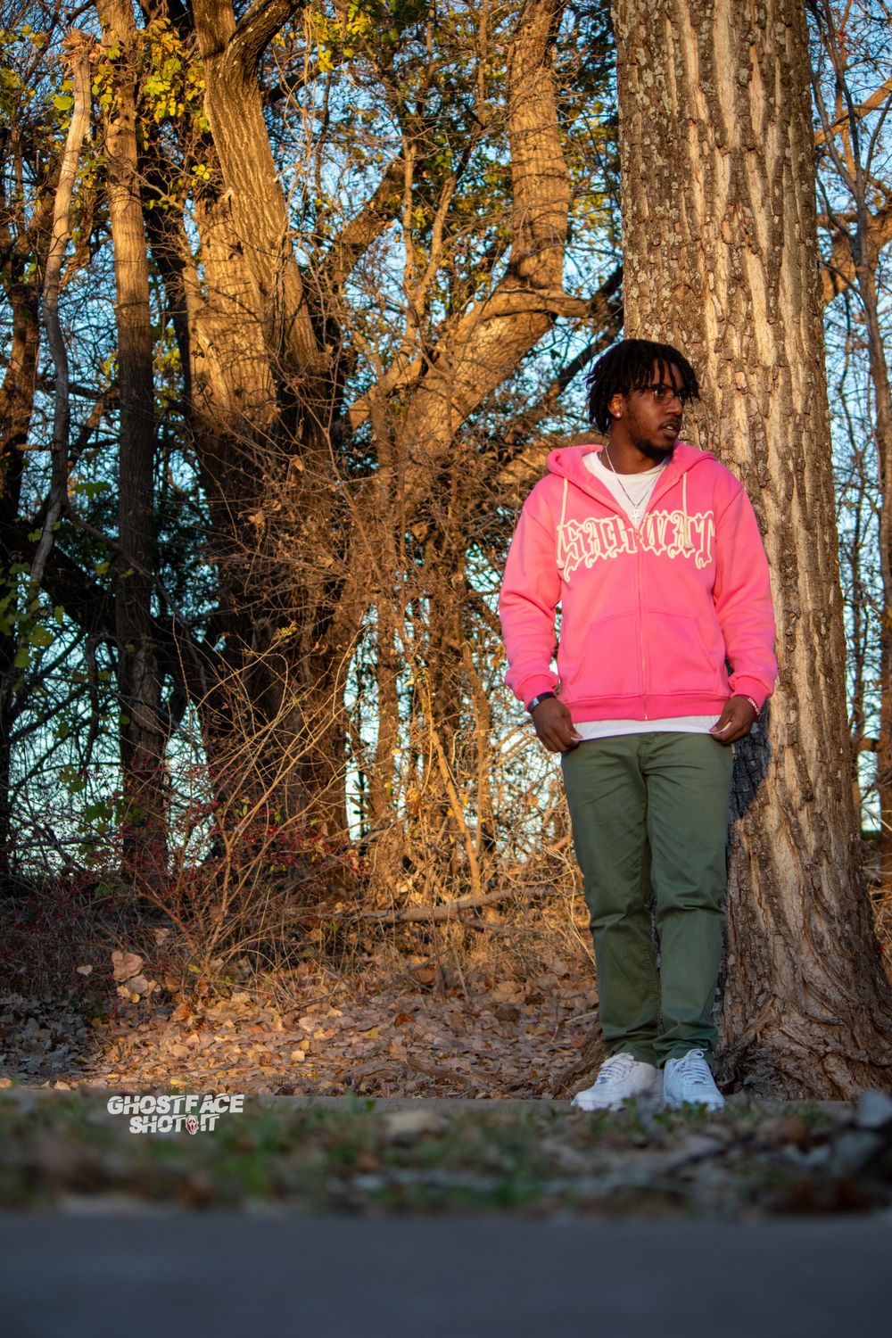 A casual outdoor portrait showing someone in a bright pink sweatshirt against sunlit trees.