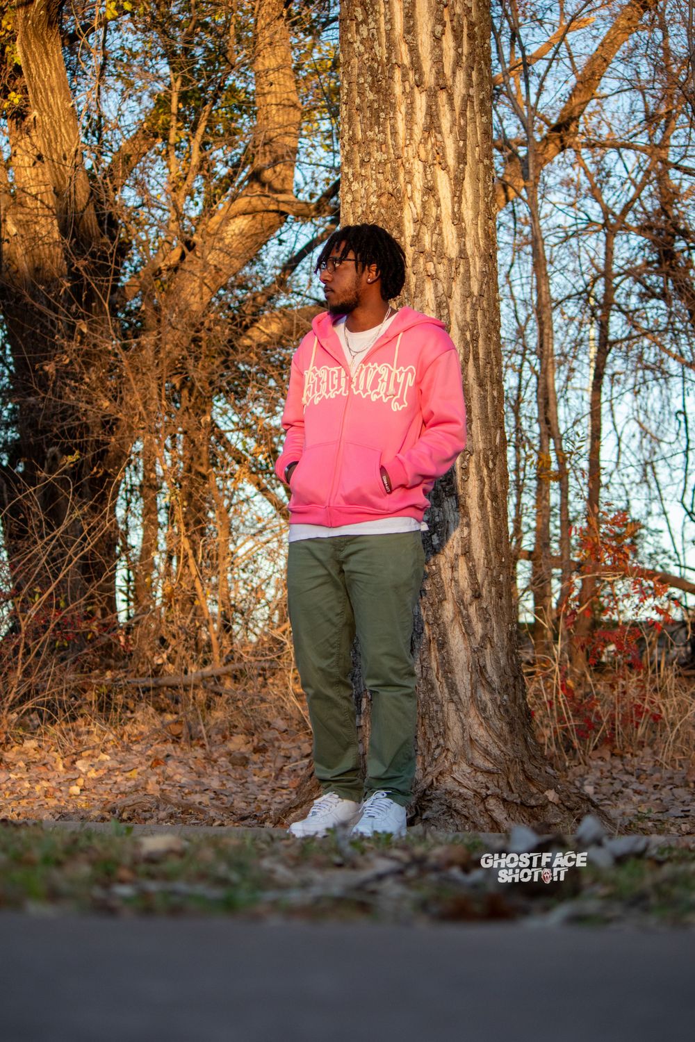 A person in a pink hoodie and olive pants stands against a tree trunk in golden afternoon light.