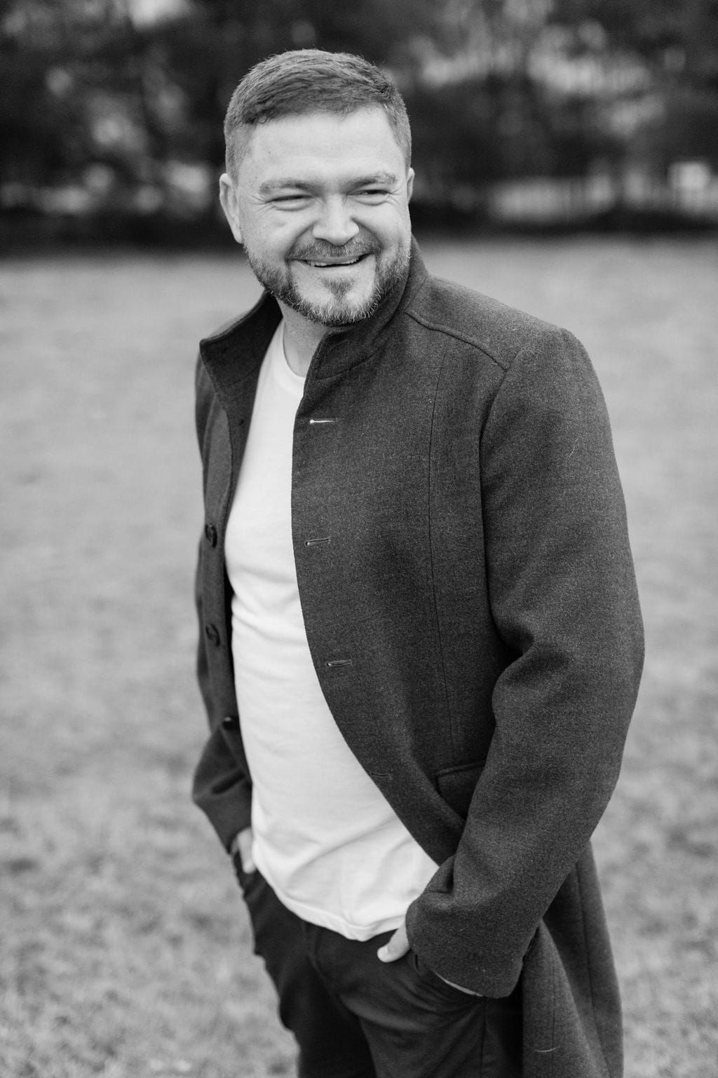 Smiling bearded man in a long coat posing outdoors in a park.