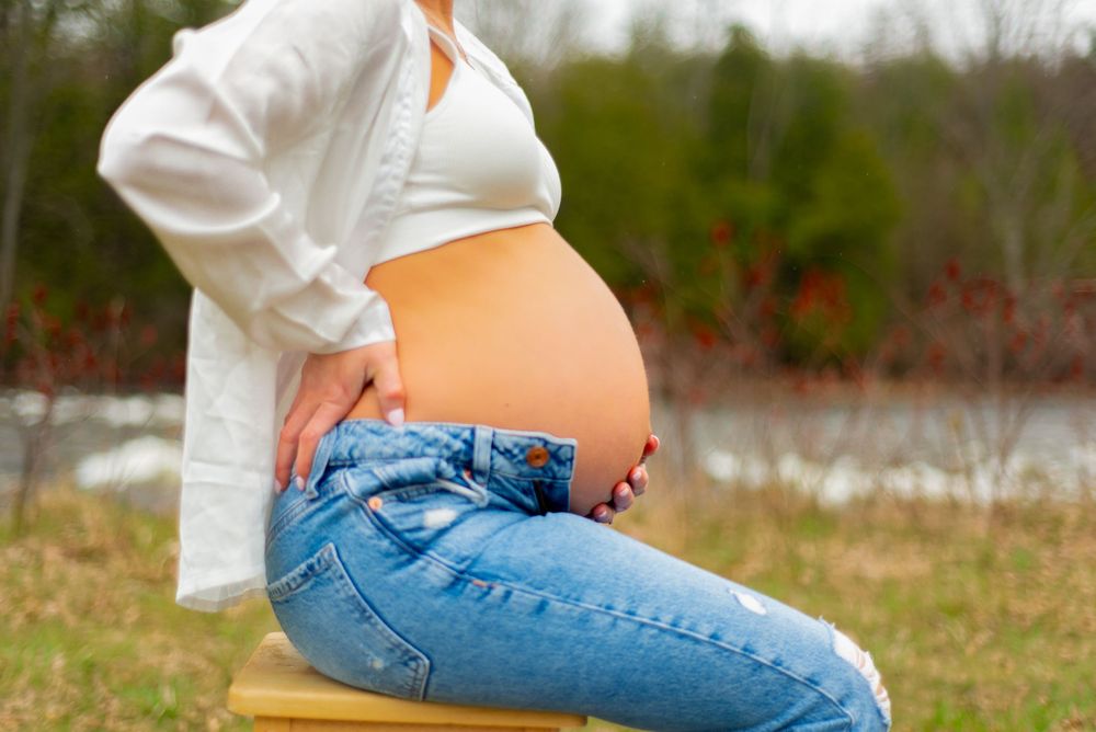 Side view of a pregnant belly in casual denim and white sweater by water.