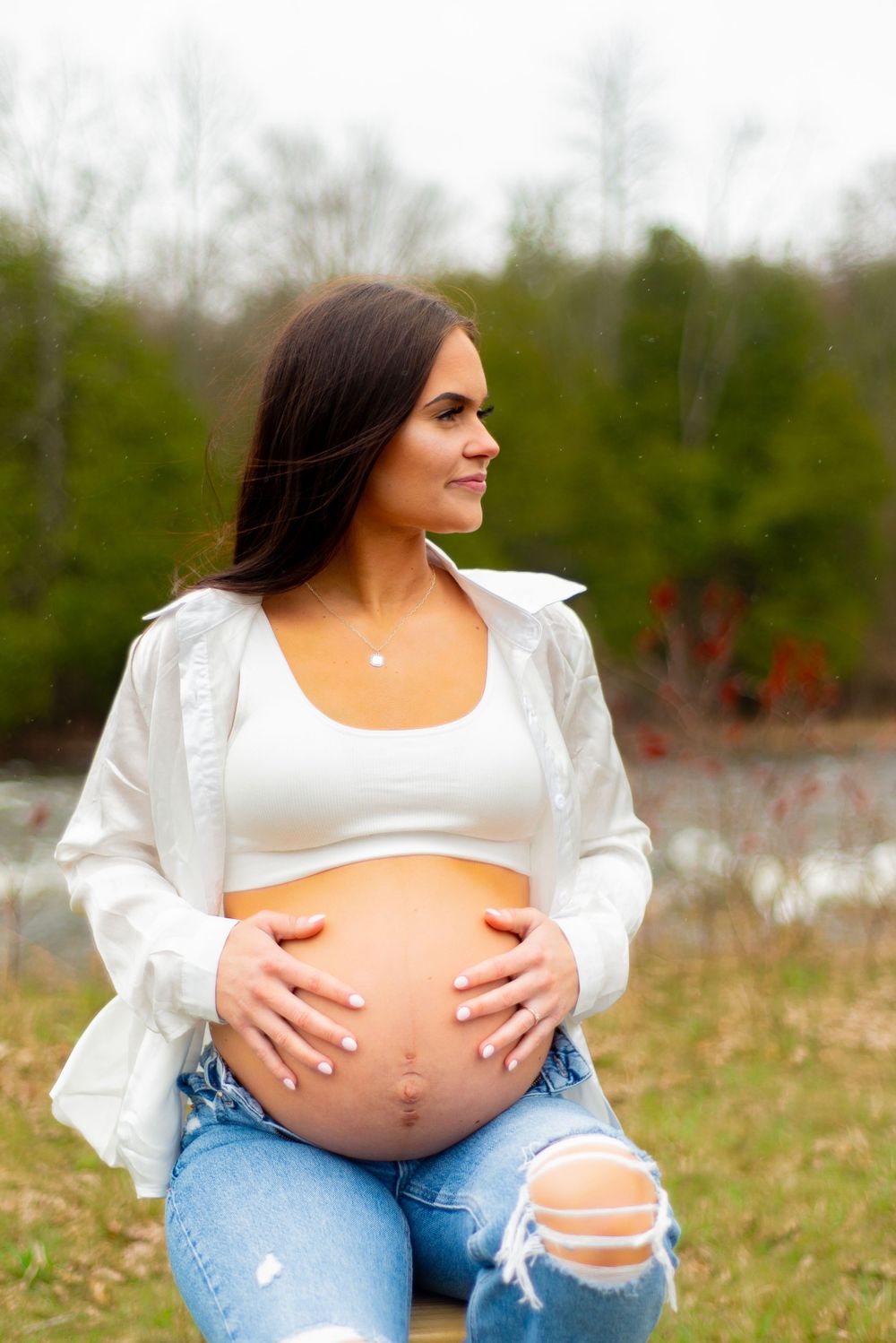 Maternity photo in ripped jeans and white crop top by a riverside setting.