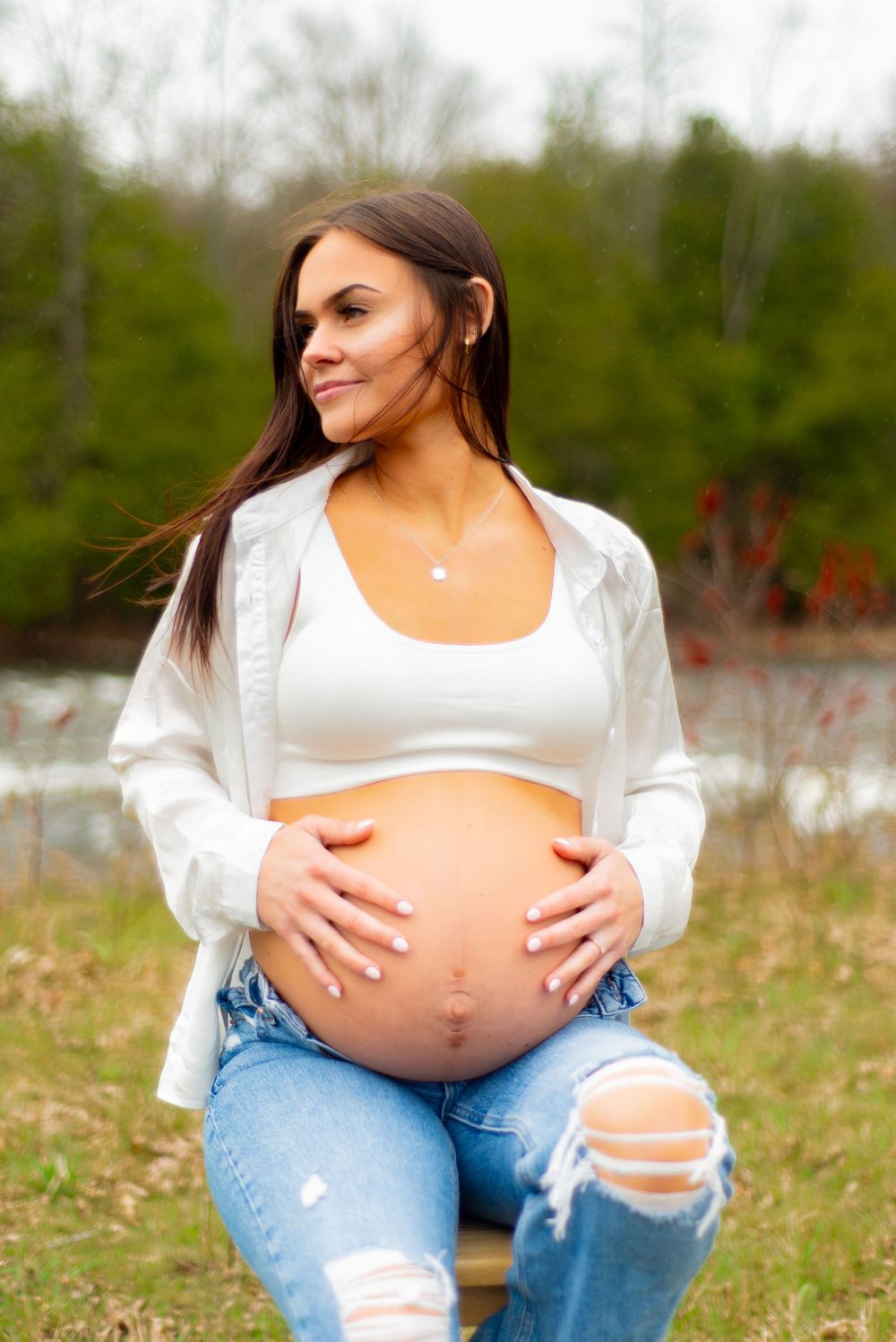 Maternity photo of pregnant person in white top and ripped jeans touching belly in outdoor nature setting.