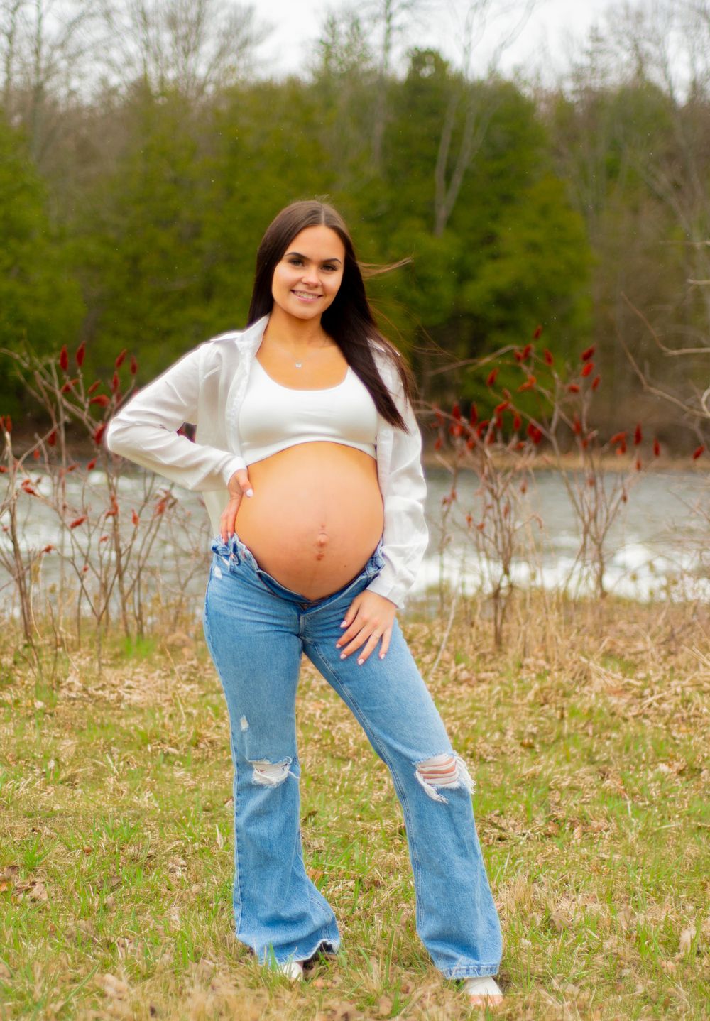 Maternity portrait showing pregnant belly while standing in casual wear against natural backdrop.