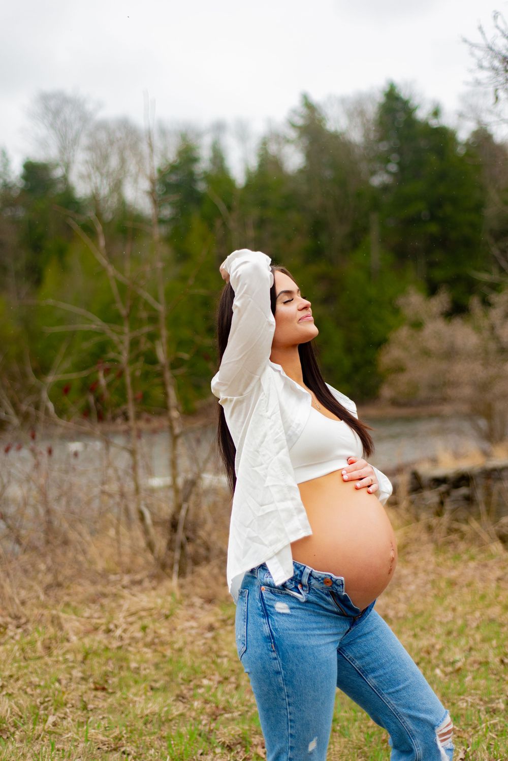 Side view of pregnant person in white shirt and jeans posing outdoors with hands raised.