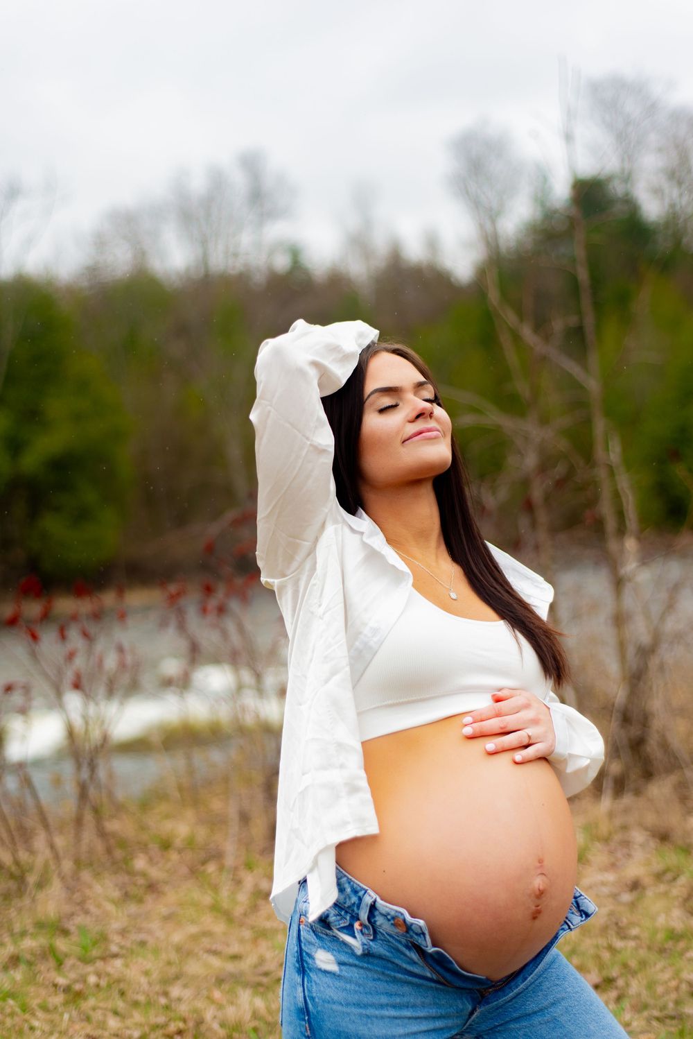 Pregnancy portrait showing bare belly while wearing white top and jeans in natural setting.