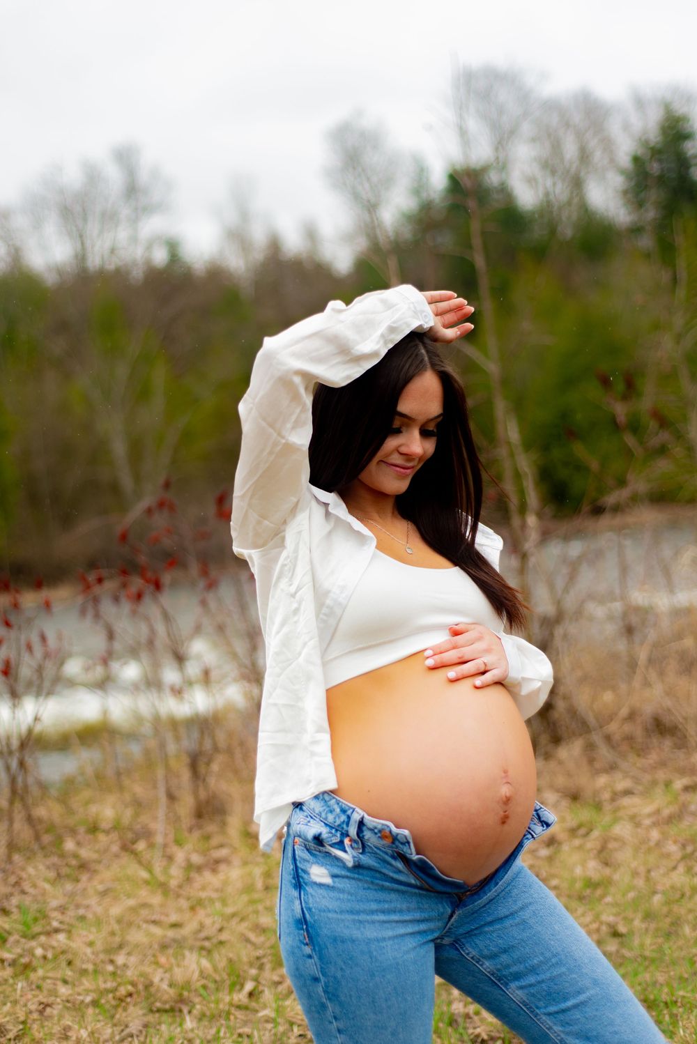 Artistic maternity photo with person in white shirt and jeans touching pregnant belly outdoors.