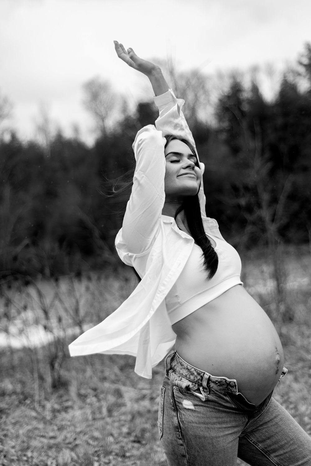 Black and white pregnancy portrait with arms raised in graceful pose outdoors.