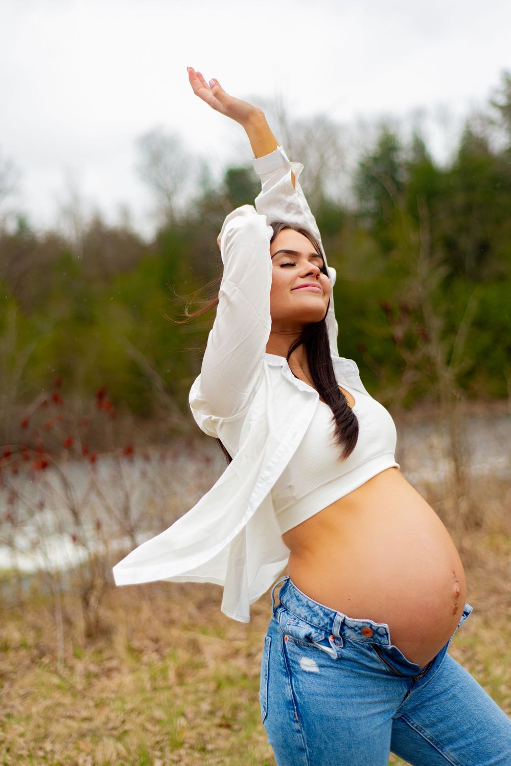 Pregnant woman in white shirt and jeans strikes joyful pose outdoors in natural setting.