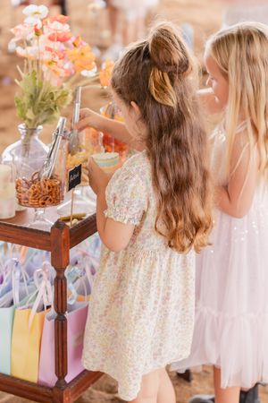 Young children in white dresses gather around a party table with pink and peach floral decorations.