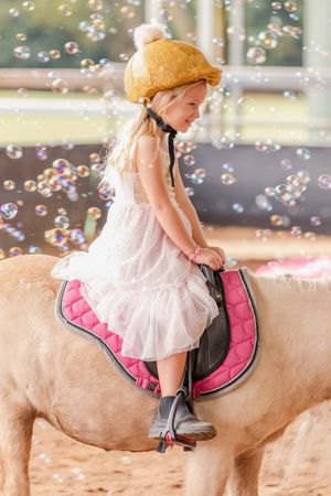 A child in a white dress riding a pony surrounded by floating bubbles.