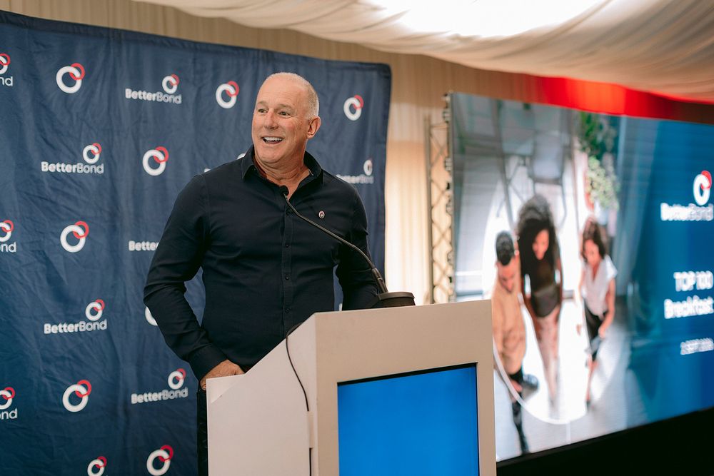 Man in black shirt speaking at BetterBond podium during corporate event with presentation screen in background.