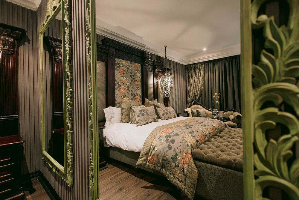 Luxurious dark hotel bedroom with ornate green carved mirror, floral bedding, four-poster bed, and dramatic moody decor.