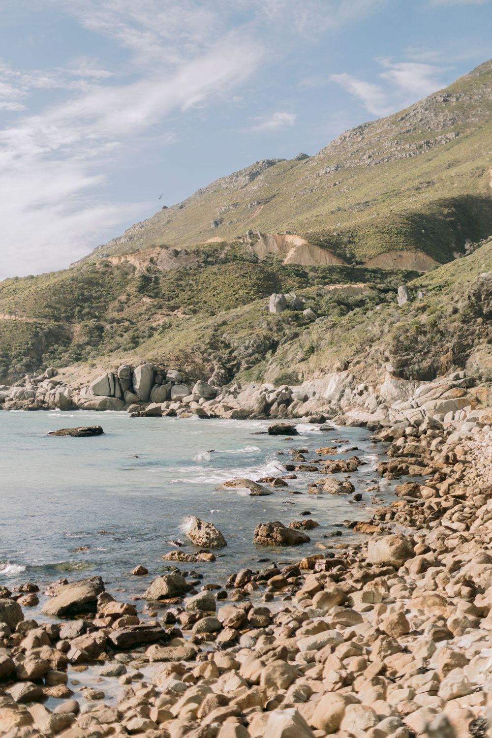 Rocky coastline with turquoise ocean waters and green mountain slopes under blue sky.