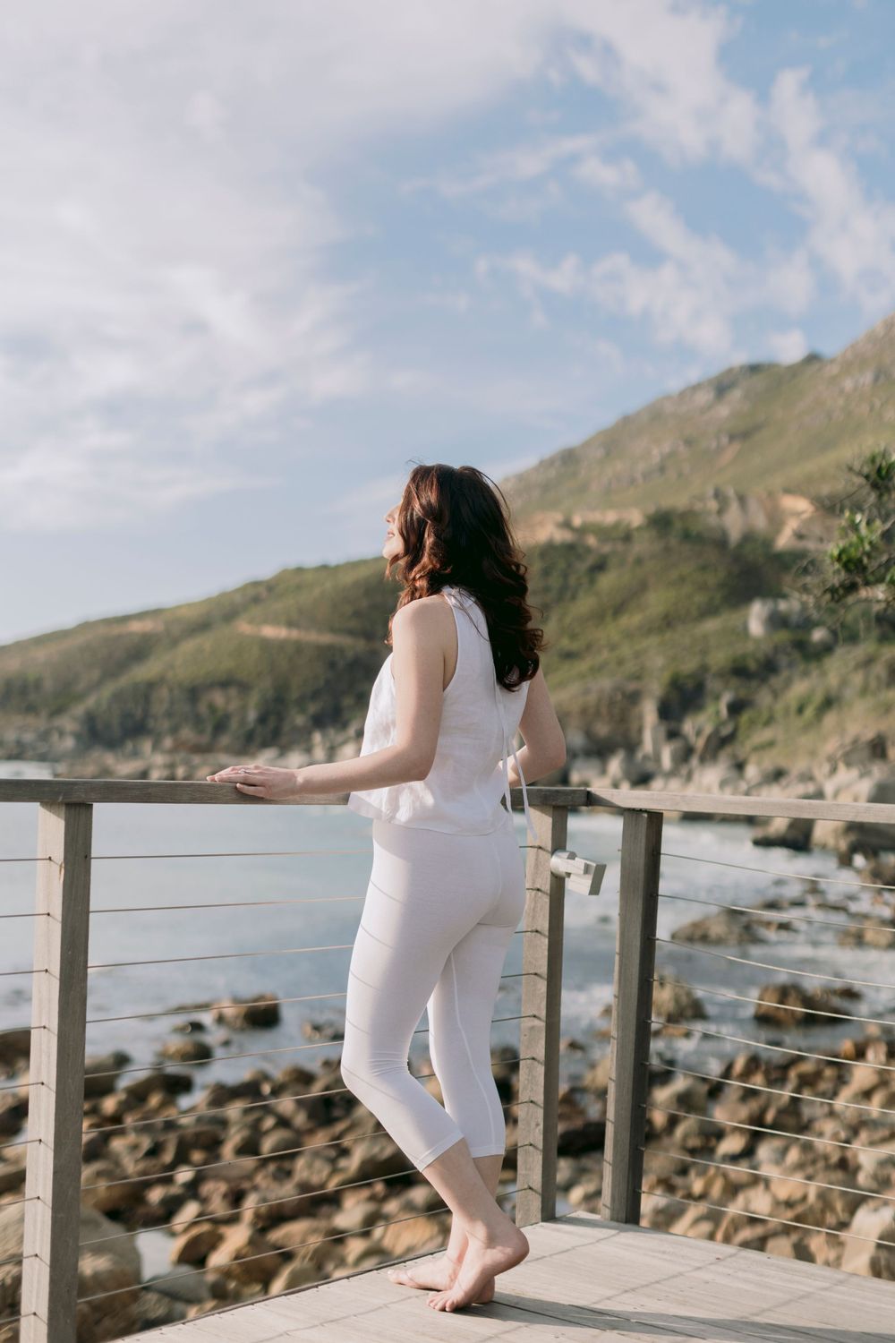 Woman in white outfit standing on wooden deck overlooking rocky coastline and green mountains.