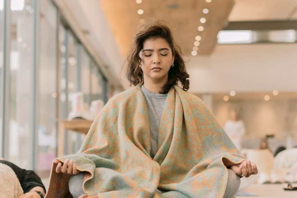 Young woman meditating in lotus pose wrapped in a colorful pastel blanket in a bright indoor studio.