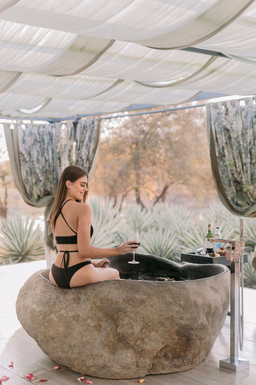 Woman in black bikini relaxing by a stone bath with red wine at luxury spa resort.