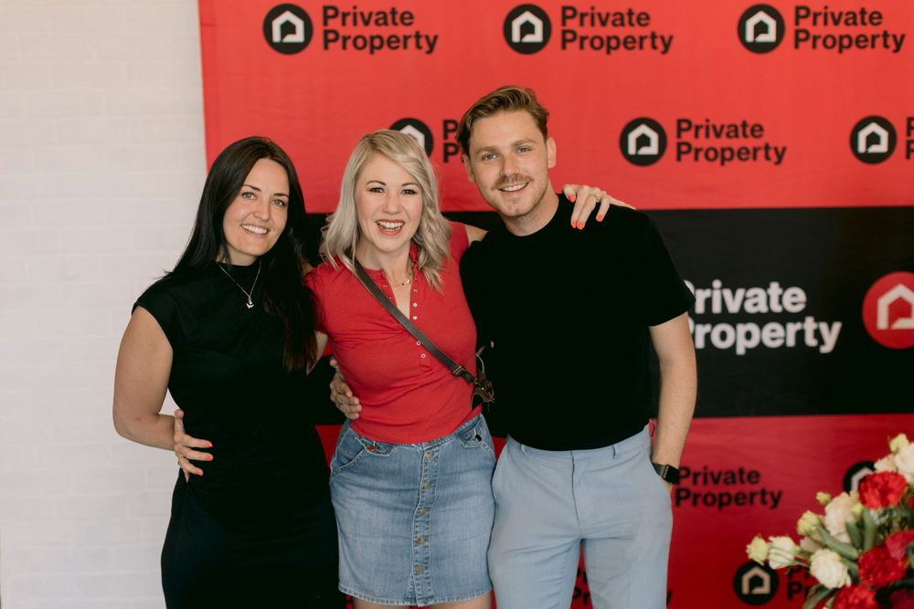 Three people smiling together in front of a Private Property branded red step-and-repeat banner.
