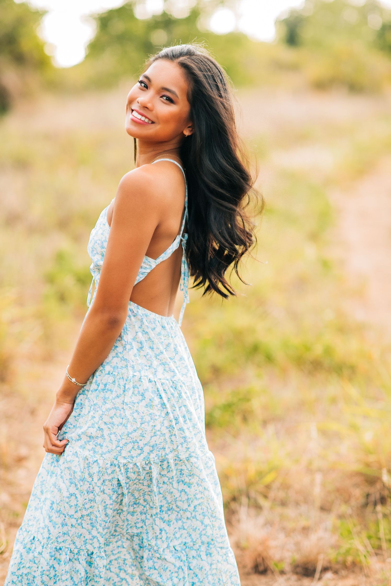 Woman in floral blue sundress poses in a sunny field with flowing dark hair.
