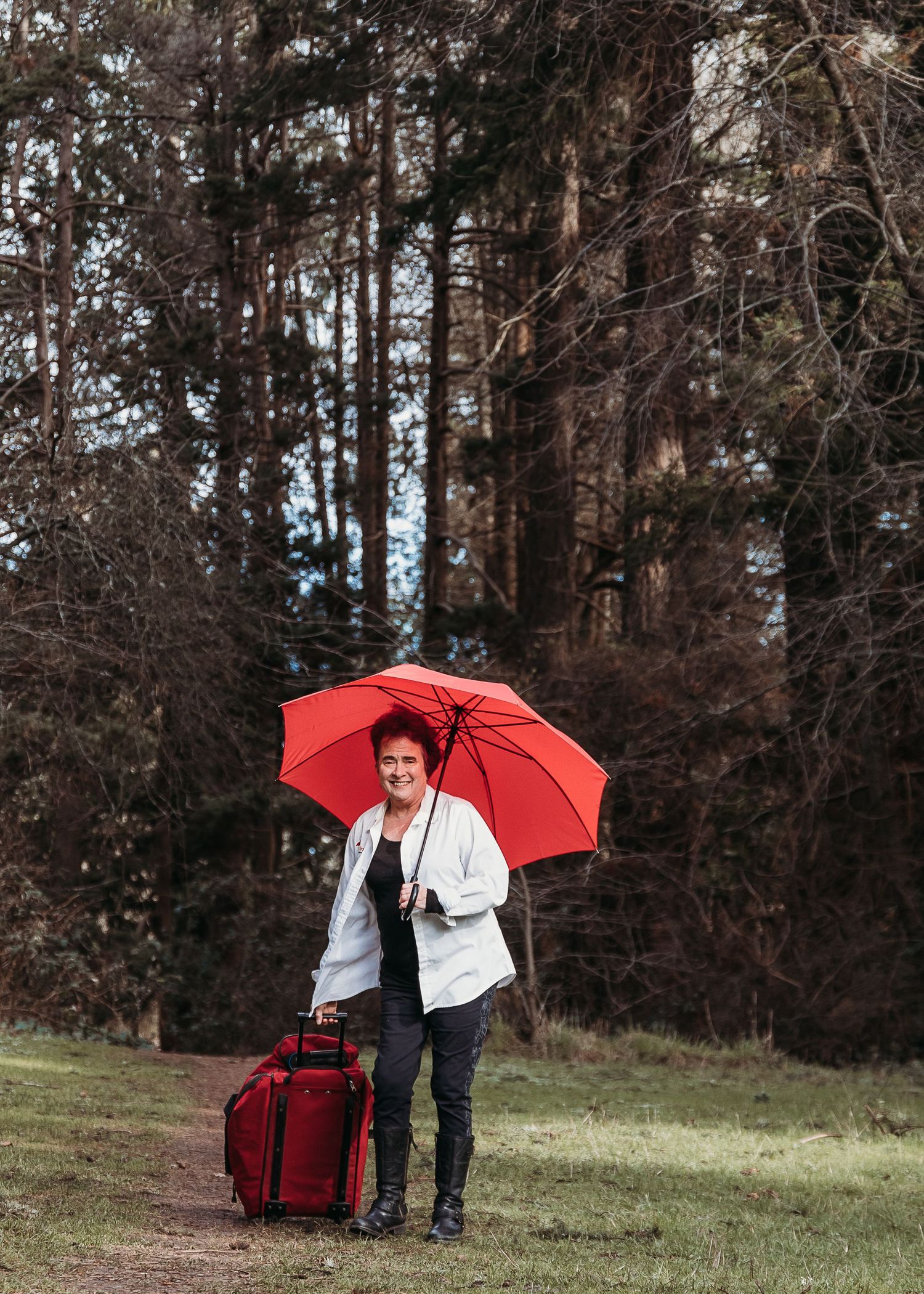 Person with red umbrella and rolling suitcase stands on forest path in casual white jacket.