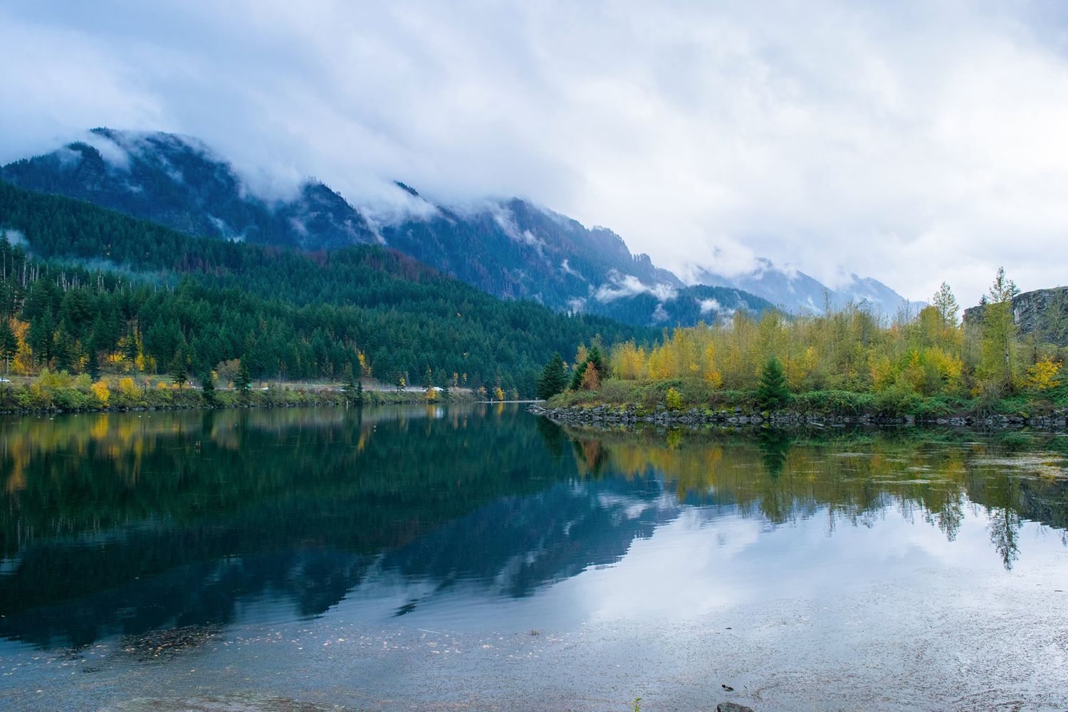 Mountain lake reflecting evergreen forest and cloudy peaks in still water.