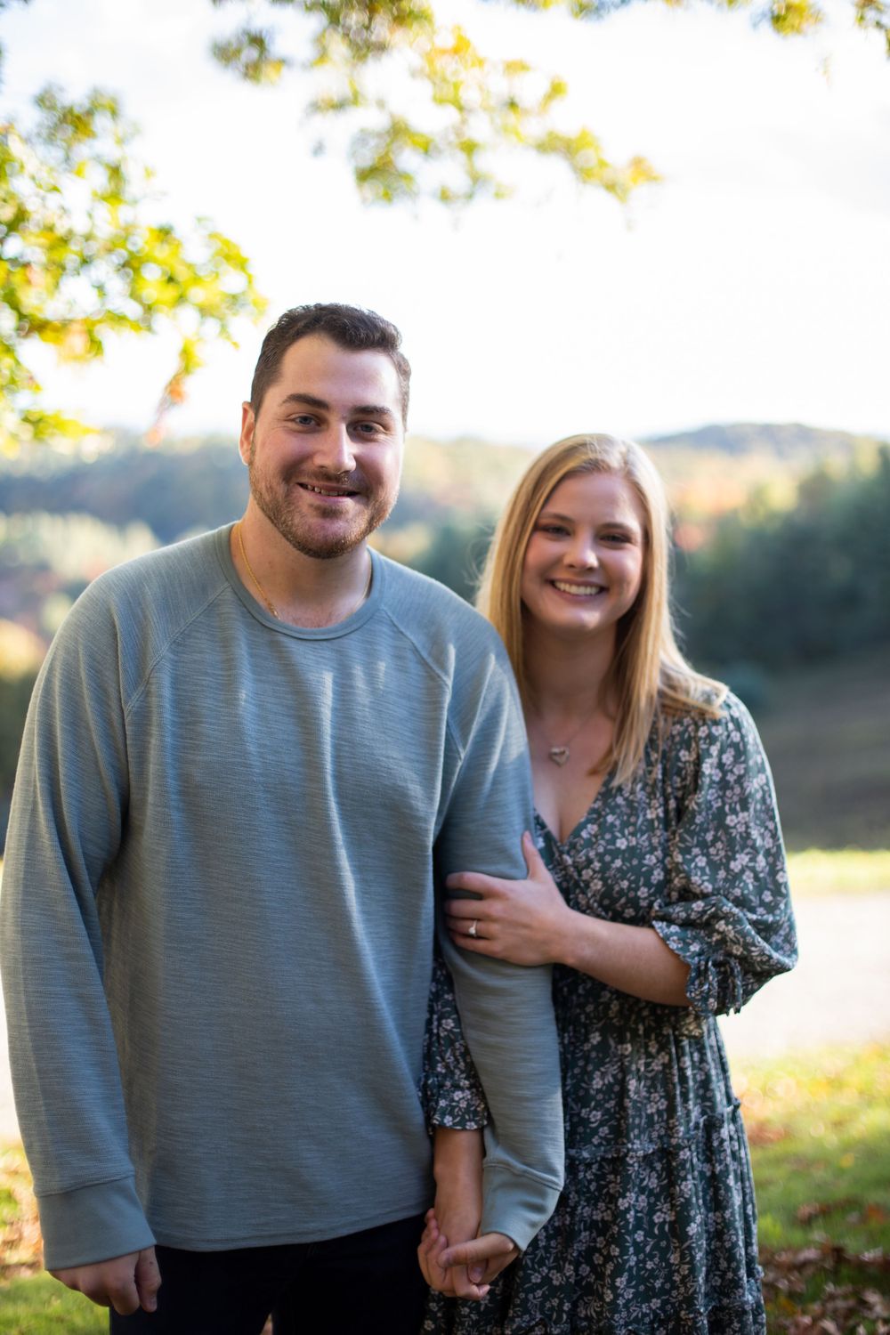 A couple poses together outdoors wearing casual attire against a scenic autumn landscape.
