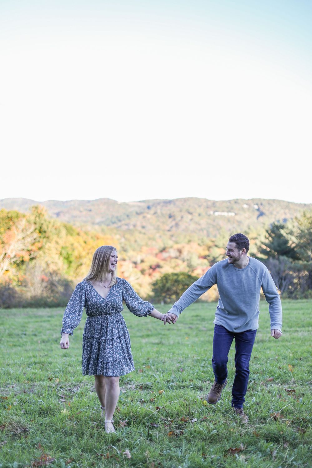 A couple holds hands while walking through a scenic hilltop meadow.