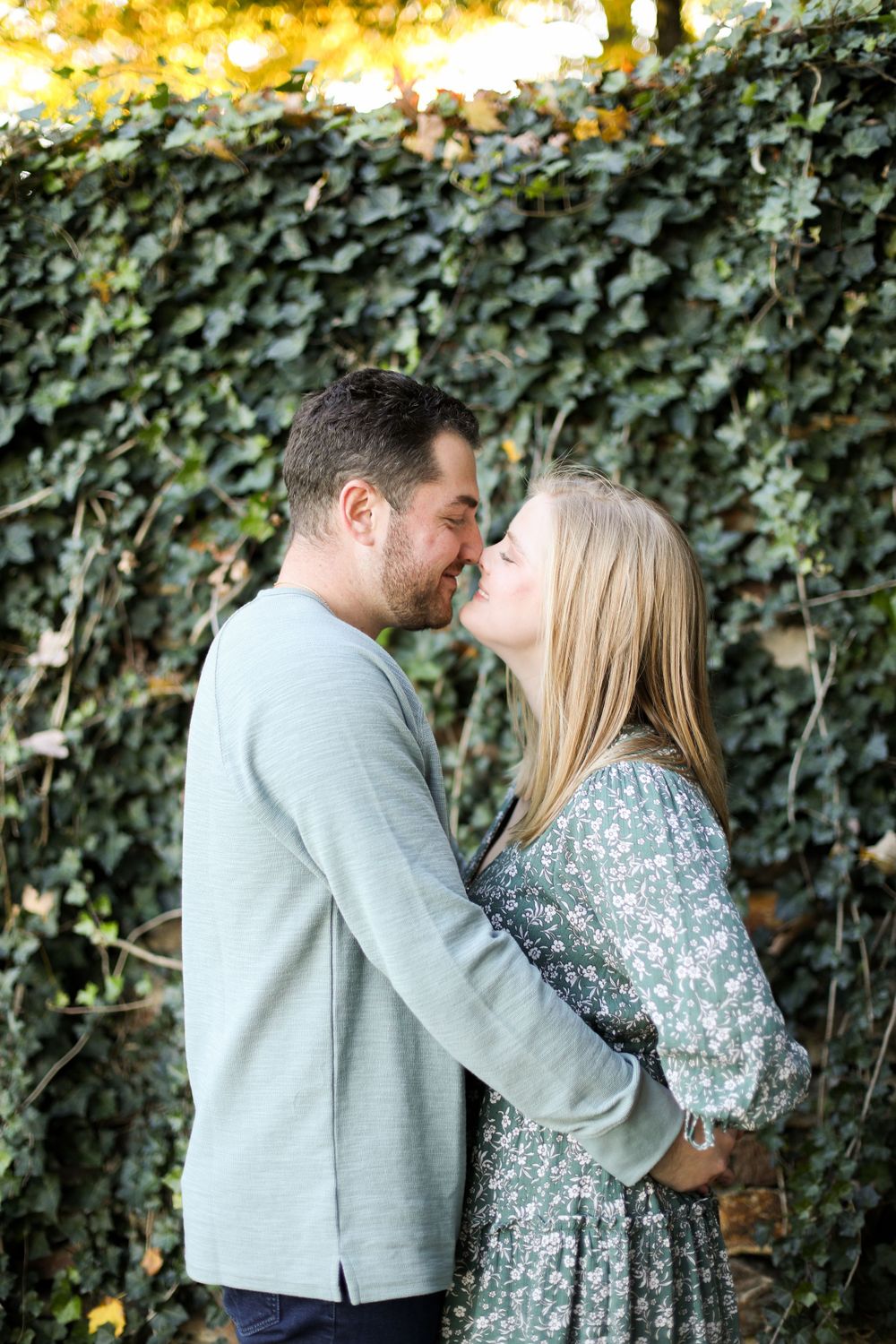 A couple shares a romantic kiss against an ivy-covered wall with golden sunlight above.
