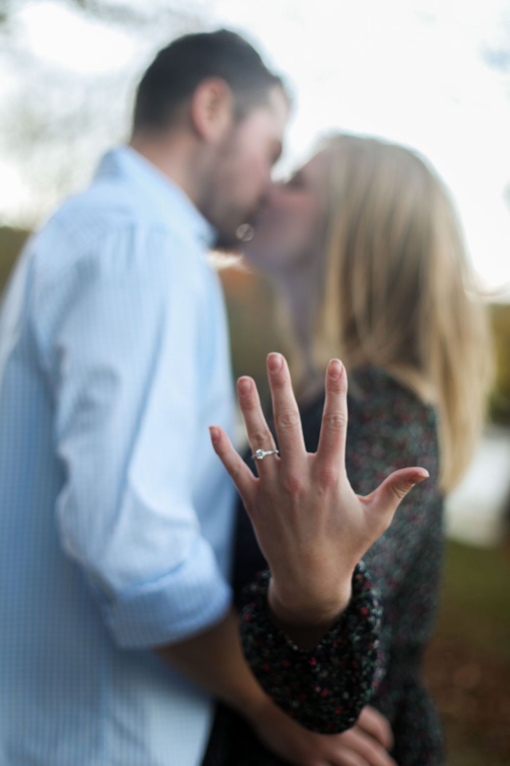 Hand displaying an engagement ring with a couple embracing in the background.