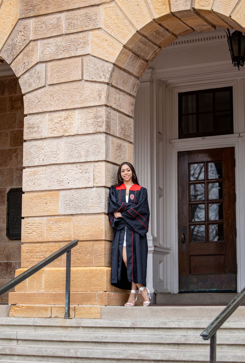 A graduate in academic regalia stands on stone steps at the entrance of a historic university building with arched stonework.